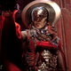 The gold armor and silky red robes from Tanta Sila shine brilliantly as she gestures in front of a red stone wall.